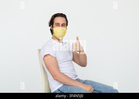 Giovane uomo caucasico felice seduto dopo il vaccino, mostra il pollice alzato, concetto di assistenza sanitaria Foto Stock
