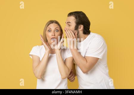 Donna e uomo sorpresi che sussurrano un segreto con espressioni, sfondo giallo Foto Stock