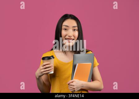 Giovane donna asiatica felice che tiene un caffè e un quaderno, sorride alla telecamera, sfondo di studio Foto Stock