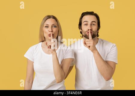 Uomo e donna che fanno il gesto del silenzio con il dito sulle labbra, sfondo giallo Foto Stock
