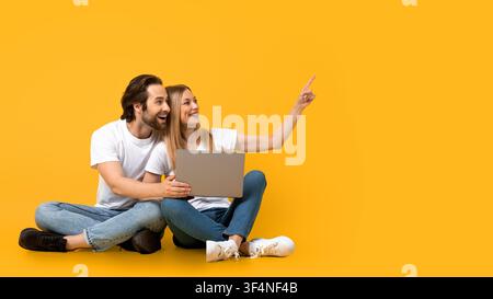 Coppia felice di giovani adulti seduti sul pavimento, laptop e indicando, espressione gioiosa Foto Stock