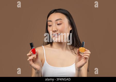 Giovane donna asiatica sorridente che tiene bottiglie di smalto per unghie rosso e giallo, concetto di bellezza Foto Stock