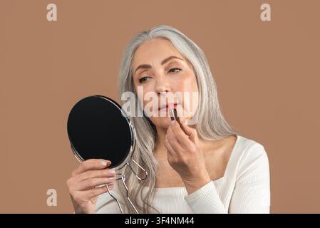 Anziana che applica il rossetto guardandosi allo specchio a casa, routine di bellezza Foto Stock