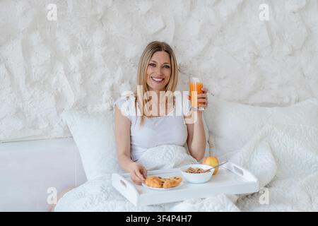 Giovane donna sorridente che gusta la colazione a letto con vassoio, pasti sani e succo fresco Foto Stock