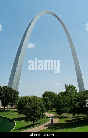 Un visitatore solitario passeggia lungo un sentiero del parco sotto l'imponente Gateway Arch in acciaio inossidabile in una limpida giornata di sole a St. Louis. Foto Stock
