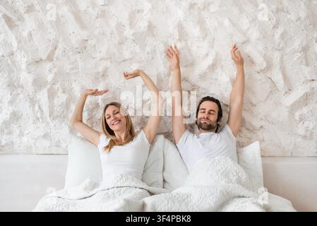 Uomo e donna caucasici felici che si allungano a letto, godendosi la mattina insieme, rilassandosi a casa Foto Stock