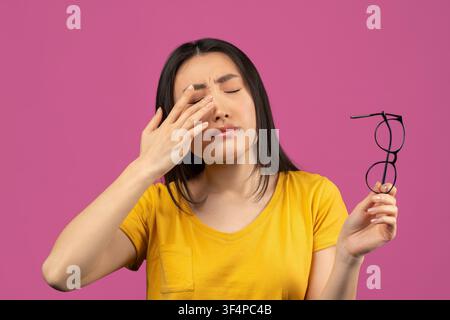 Giovane donna asiatica stressata che si strofina la fronte mentre tiene gli occhiali, sfondo rosa Foto Stock