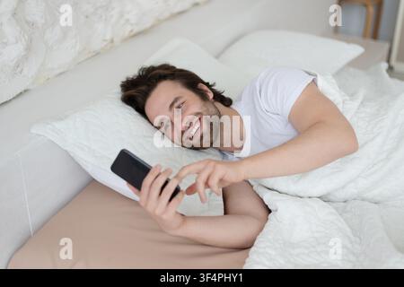 Giovane uomo caucasico felice sdraiato a letto, che usa lo smartphone al mattino, concetto di benessere Foto Stock