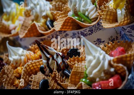 Un primo piano mostra una serie di coni di waffle ripieni di gelato morbido bianco e varie guarnizioni come sciroppo di cioccolato, pezzi di biscotto, verde Foto Stock