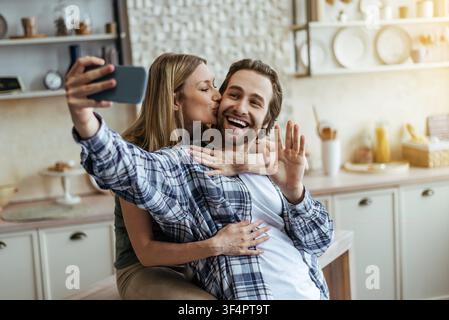 Giovane coppia felice che fa un selfie in cucina, donna che bacia l'uomo, sorridendo insieme Foto Stock