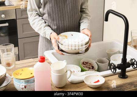 Persona in grembiule che tiene una pila di stoviglie sporche sopra un lavandino da cucina pieno di altri piatti usati Foto Stock
