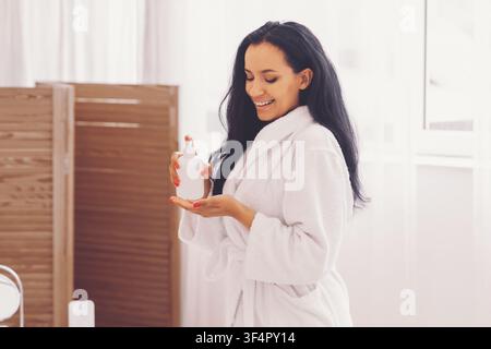 Giovane donna felice che applica siero per la cura della pelle in accappatoio bianco a casa, concetto di benessere Foto Stock
