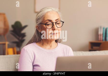 Donna anziana con capelli grigi che usa un laptop a casa, concentrata sullo schermo, rilassata Foto Stock