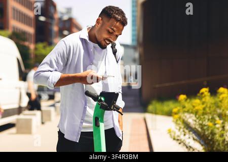 Giovane uomo nero felice che usa uno smartphone mentre è in piedi accanto a uno scooter elettrico, ambientazione urbana Foto Stock