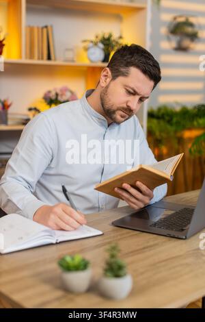 Giovane uomo sul tavolo di casa studia con laptop e libro durante la lezione online, scrive appunti e gira la pagina. Adulto sul tavolo di casa impara da un corso a distanza in una tranquilla stanza di casa con una routine di studio concentrata. Foto Stock