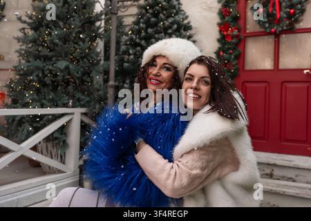 Amici in vacanza invernali che si abbracciano all'aperto durante la nevicata, indossando cappotti festivi in finta pelliccia vicino all'ingresso di una casa decorata Foto Stock
