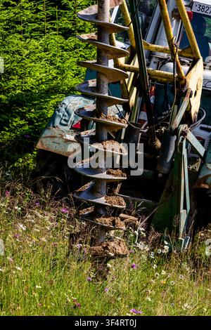 Una trivella robusta ricoperta di terra fresca pende da un trattore accanto a un prato fiorito. Le lame rotanti e le braccia idrauliche mostrano un lavoro rurale pratico in pr Foto Stock