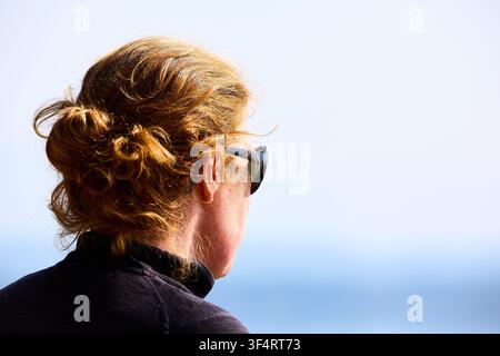 Una donna dai capelli rossi con occhiali da sole e una maglietta sportiva che guarda un lago in una giornata luminosa e soleggiata Foto Stock