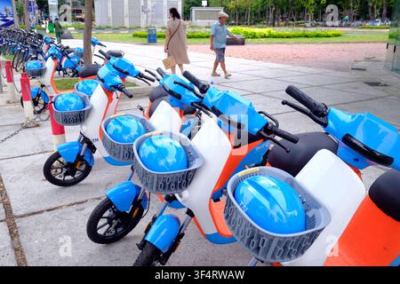 Una fila di scooter elettrici blu e bianchi, ognuno con un casco blu nel cestino, attende gli utenti in un parcheggio urbano. Ho Chi Minh City, Vietnam. Foto Stock