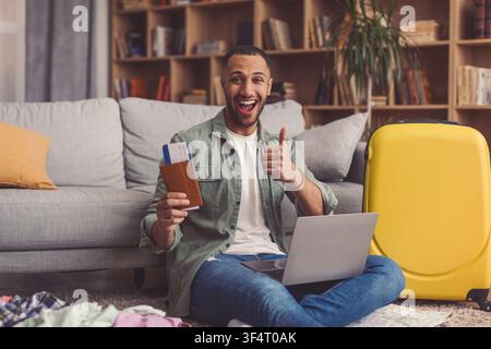 Uomo felice che usa un laptop tenendo un passaporto e dei bagagli nel soggiorno, concetto di viaggio Foto Stock