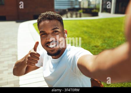 Giovane uomo sorridente con auricolari che dà il pollice alzato all'aperto nel parco cittadino, concetto di stile di vita Foto Stock