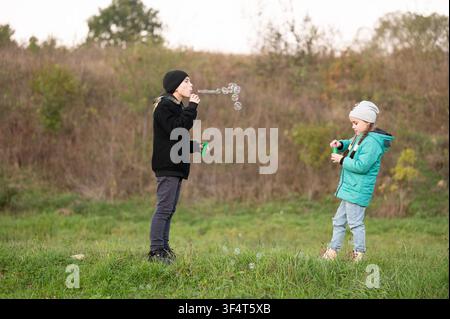 Due bambine soffiano felicemente bolle di sapone in un pomeriggio sereno. Una bambina con una giacca nera soffia bolle mentre un’altra con un cappotto verde acqua gioca, entrambe si divertono Foto Stock