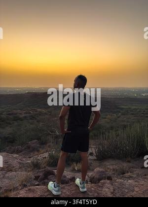Silhouette di un uomo solitario in piedi sulla cima di una collina durante un tramonto colorato con le luci della città Foto Stock