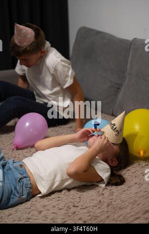 Due bambini partecipano a festività di compleanno giocose al chiuso, con uno sdraiato su un tappeto morbido e l'altro seduto nelle vicinanze, circondato da palloncini vibranti Foto Stock