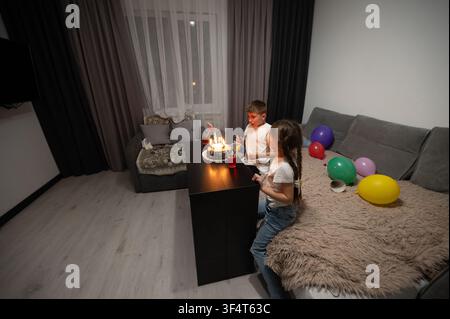 Due bambini accendono le candeline di compleanno su una torta, circondati da palloncini vibranti e da un'atmosfera calda e accogliente, creando ricordi preziosi durante un gioioso Foto Stock
