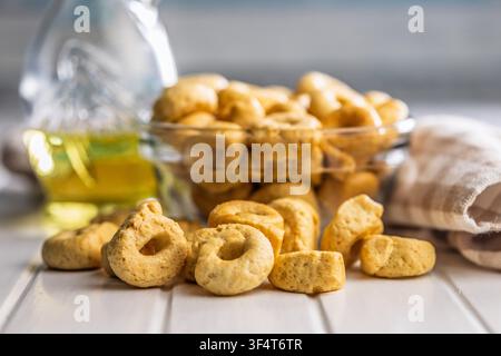 Cracker taralli dorati a forma di anello ammucchiati in una ciotola di vetro accanto a una bottiglia di olio d'oliva e un canovaccio piegato, che trasmettono una rustica e croccante Medit Foto Stock