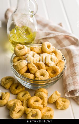 Cracker taralli dorati a forma di anello ammucchiati in una ciotola di vetro accanto a una bottiglia di olio d'oliva e un canovaccio piegato, che trasmettono una rustica e croccante Medit Foto Stock