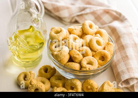 Cracker taralli dorati a forma di anello ammucchiati in una ciotola di vetro accanto a una bottiglia di olio d'oliva e un canovaccio piegato, che trasmettono una rustica e croccante Medit Foto Stock