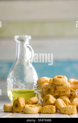 Cracker taralli dorati a forma di anello ammucchiati in una ciotola di vetro accanto a una bottiglia di olio d'oliva e un canovaccio piegato, che trasmettono una rustica e croccante Medit Foto Stock