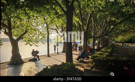 Le persone si godono una passeggiata lungo un sentiero ombreggiato dagli alberi sulla riva, suonatori di accordion seduti sulla riva soleggiata, persone sulle panchine, passeggiata di Greenwich, Tege Foto Stock