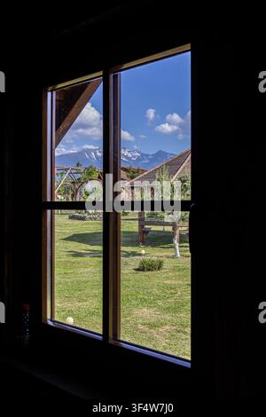 Un colpo prospettico verticale dall'interno che guarda attraverso una finestra con cornice in legno scuro su un paesaggio luminoso e soleggiato con erba verde e montagne lontane Foto Stock
