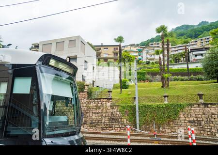Caslano - Svizzera - 12 maggio 2026: Vista ravvicinata della cabina di guida di un treno con binari ferroviari, segnali di sicurezza e un villaggio collinare con palme nel Foto Stock