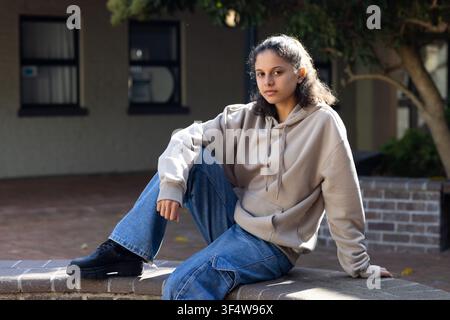 Adolescente afroamericano seduto su un basso muro di mattoni in un cortile, indossa una felpa con cappuccio beige e stivali Foto Stock