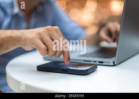 Scatto mirato di mani maschili che fanno multitasking. L'indice destro tocca lo schermo di uno smartphone mentre la mano sinistra lavora su un laptop. L'uomo d'affari tasta una notifica Foto Stock