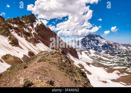 Escursionista solitario si trova in cima a una cresta montuosa frastagliata. La neve cade dolcemente lungo i pendii vulcanici. Il cielo esplode di blu con soffici nuvole bianche. Il picco lontano brilla wi Foto Stock