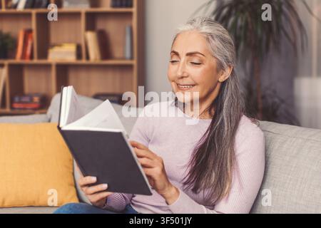 Donna anziana felice che legge un libro sul divano a casa, rilassandosi e godendosi il tempo Foto Stock