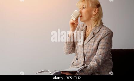 Donna anziana che si rilassa durante una pausa in ufficio, godendosi una tazza di caffè calda mentre legge una rivista, trovando un momento di calma e contemplazione in Foto Stock