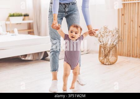 Bambina felice che tiene le mani di una donna caucasica mentre impara a stare in piedi a casa Foto Stock