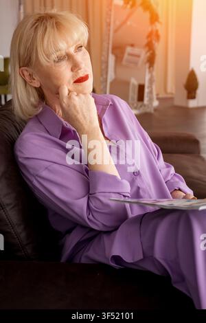 Donna bionda anziana in un abito viola seduta su un divano a casa, pensa e riflette sulla vita, mostrando pensiero e solitudine Foto Stock