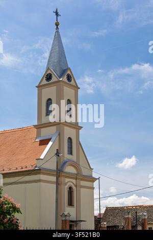 Una chiesa di villaggio storica presenta un alto campanile con una torre dell'orologio e una croce di metallo sopra un tetto di tegole rosse, sullo sfondo di un cielo blu brillante con delicatezza Foto Stock