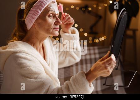 Donna matura in accappatoio che si prende cura della sua pelle, massaggia il viso applicando schiuma di pulizia con una spazzola in silicone guardandosi allo specchio. Idratazione cutanea, anti agi Foto Stock