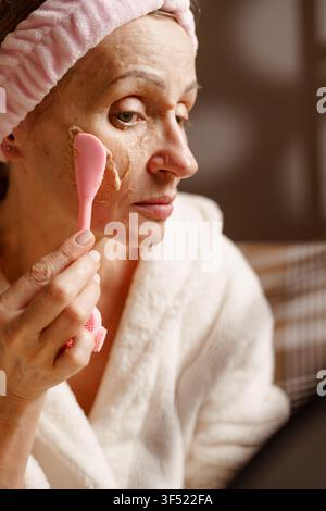 Concetto di cura della pelle matura. Donna anziana con fascia per capelli che esegue un massaggio facciale con scrub organico, applica crema scrub, peeling della pelle del viso Foto Stock
