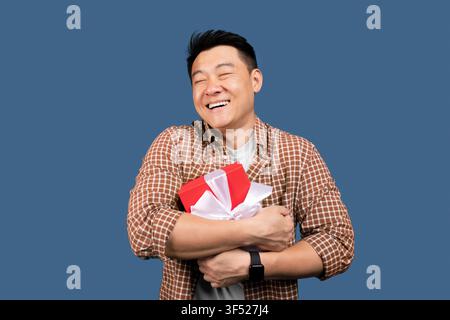 Uomo asiatico felice che tiene una scatola regalo con nastro, sorridente, sfondo blu, concetto di celebrazione Foto Stock