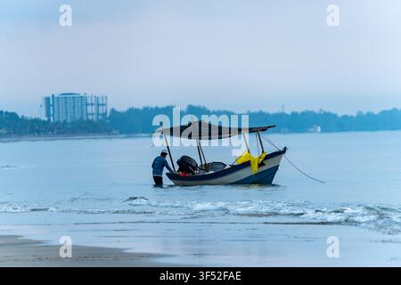 Kuantan, Malesia - 18 aprile 2026 - Un pescatore si trova nell'acqua bassa, curando la sua piccola barca sulla costa. Il mare calmo e la luce soffusa Foto Stock
