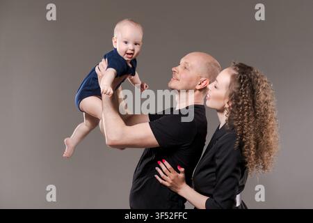 Una famiglia felice che ride mentre il padre solleva il loro bambino e la madre sorride su uno sfondo grigio, un momento di gioia condivisa con spazio per il testo Foto Stock