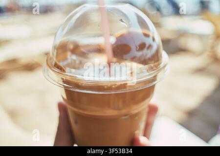 Primo piano di un caffè freddo rinfrescante in una tazza di plastica trasparente con una cannuccia rosa, tenuto in mano su uno sfondo sfocato. Foto Stock